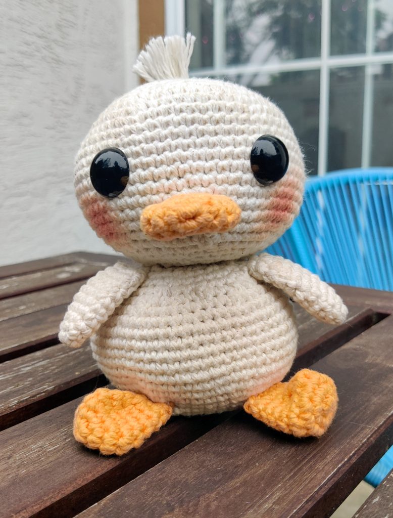 A crocheted duck plush with large black eyes and orange beak and feet sits on a wooden table outdoors.