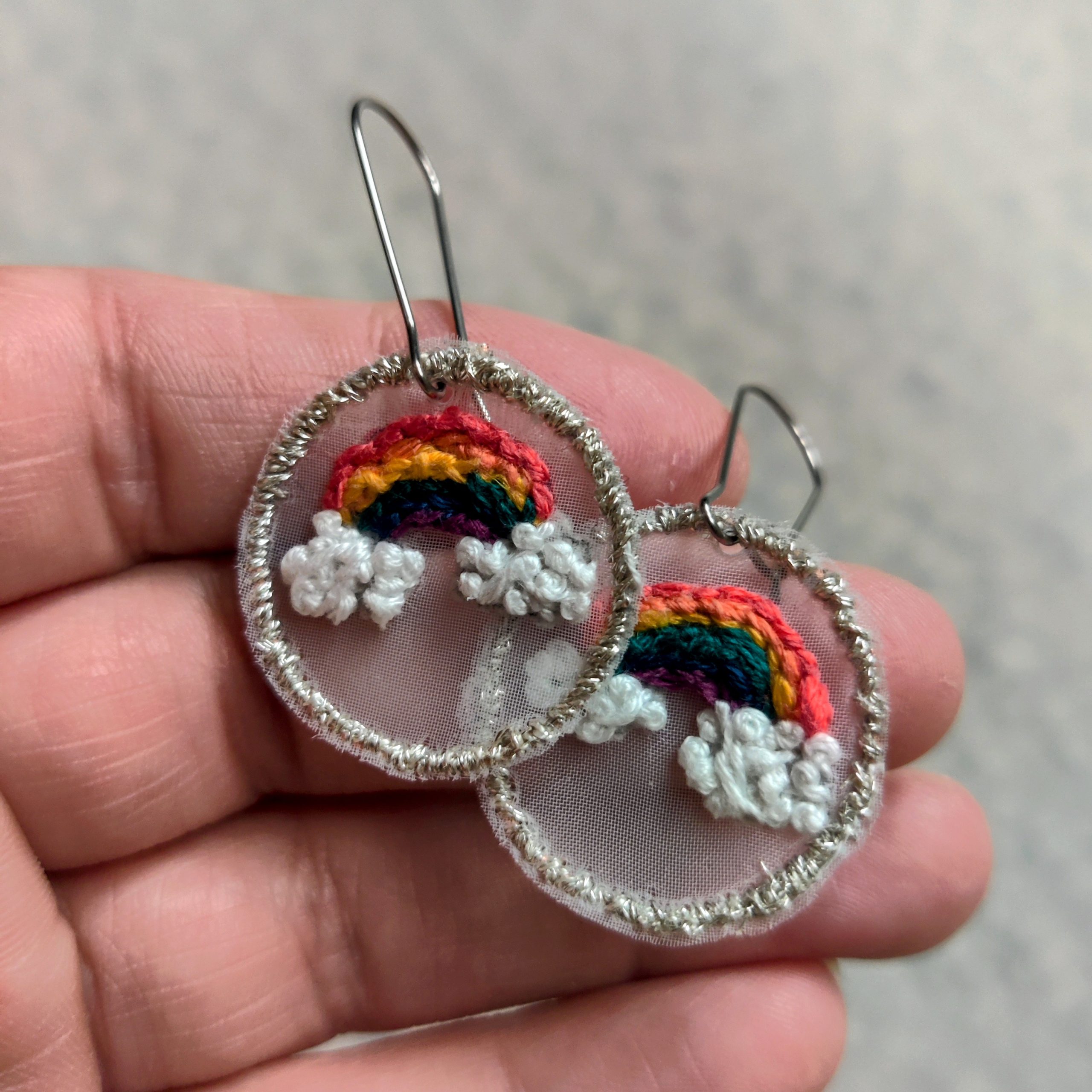Close-up of two handmade embroidered earrings featuring small rainbow arches with white cloud details, held between fingers.