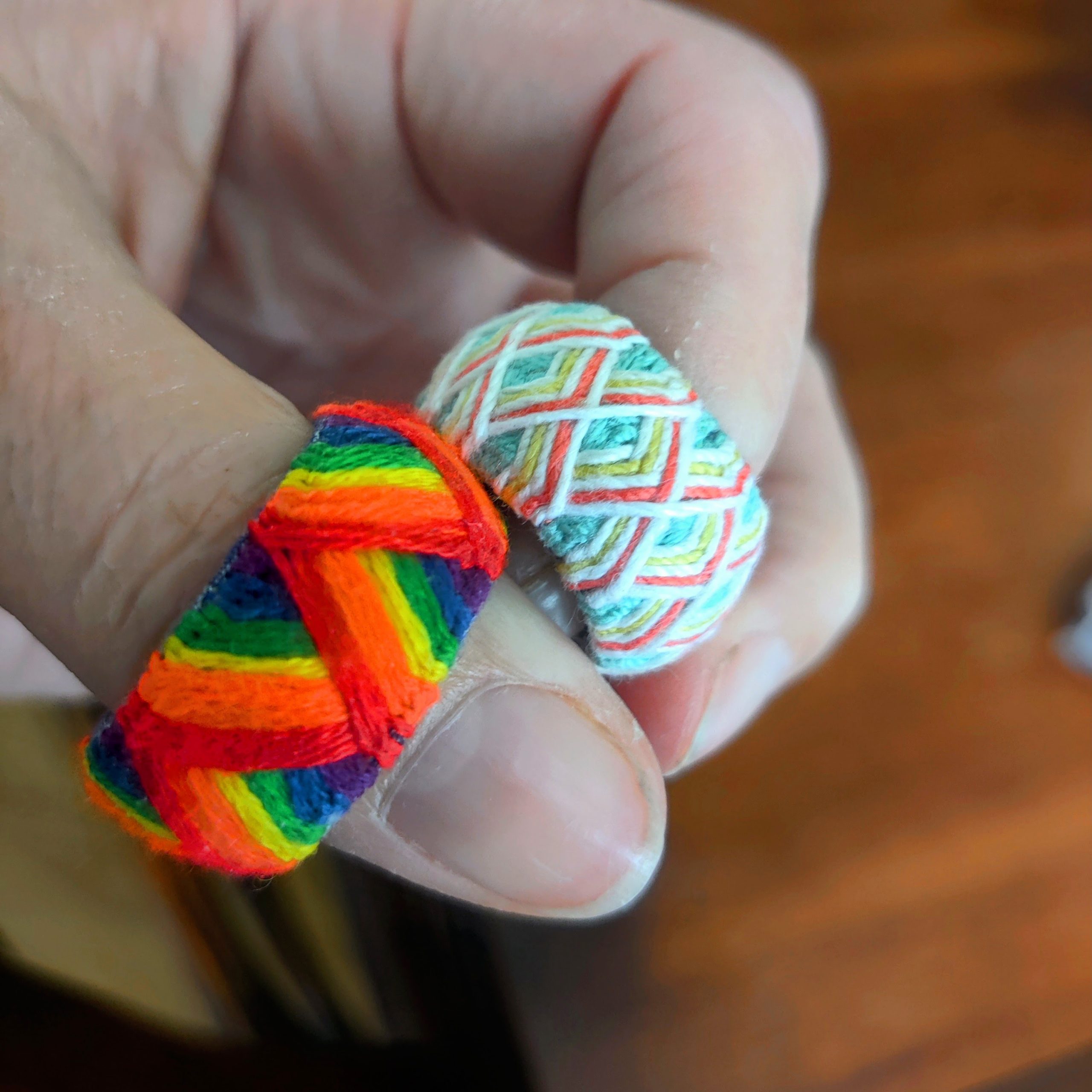 Fingers held together displaying two fiber-wrapped rings: a bold rainbow-striped ring and a lighter ring with intersecting pastel lines.