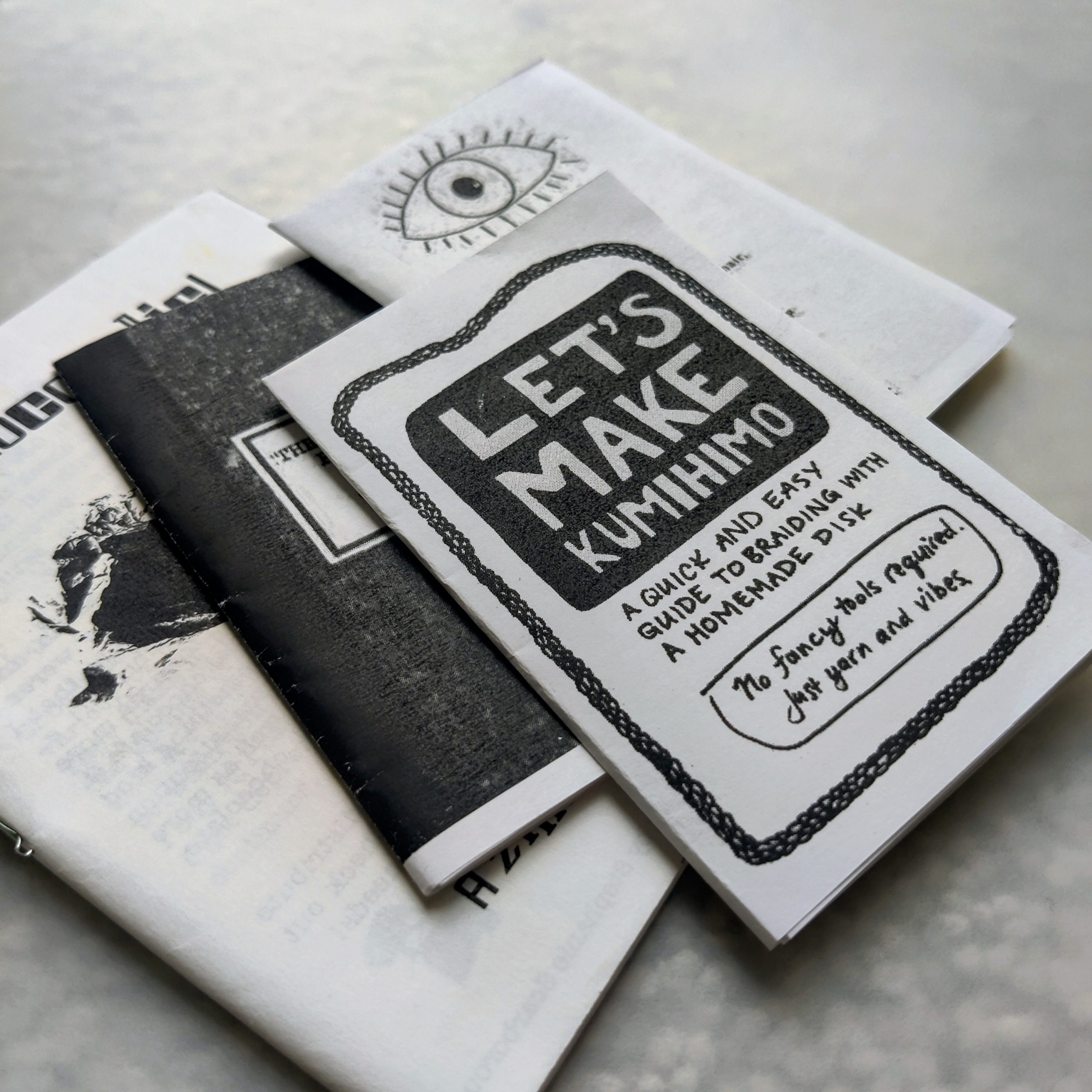 A small stack of black-and-white zines or booklets, with the top cover reading “Let’s Make Kumihimo,” photographed on a light surface.