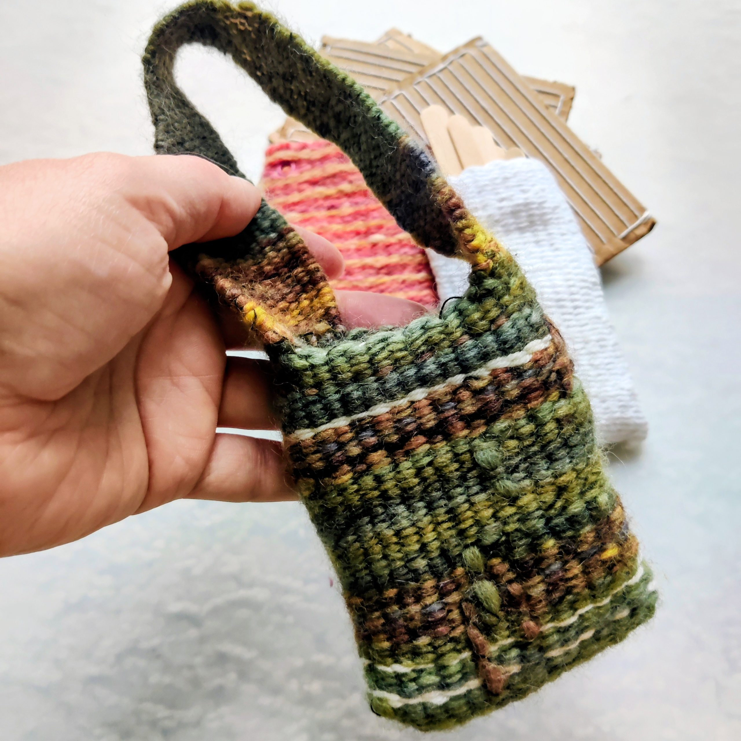 Hand holding a small, handmade woven pouch with a loop handle, featuring green and brown variegated yarn; craft materials lie blurred in the background.